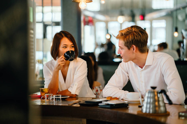 Chinese Asian woman is having coffee with a young Caucasian man. They are catching up casually in the day in a cafe or coworking space and are smiling as they talk comfortably. - Φωτογραφία, εικόνα