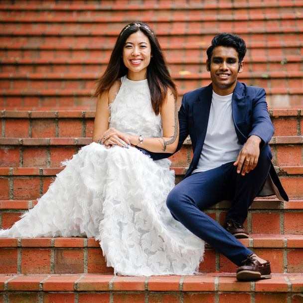 mixed race couple sitting on orange brick steps in a park in Singapore. They are newly engaged and taking photographs for their wedding as is custom. He is an Indian man, and she a Chinese woman. - Fotografie, Obrázek