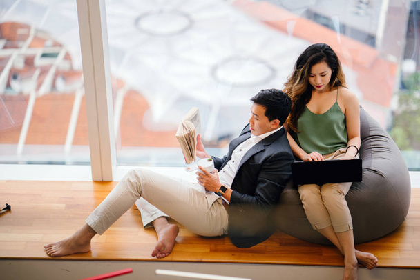 A Chinese Asian couple unwind over the weekend in their apartment. An attractive woman is working on her laptop on a beanbag and her boyfriend is reading a book and leaning against her. - Фото, изображение