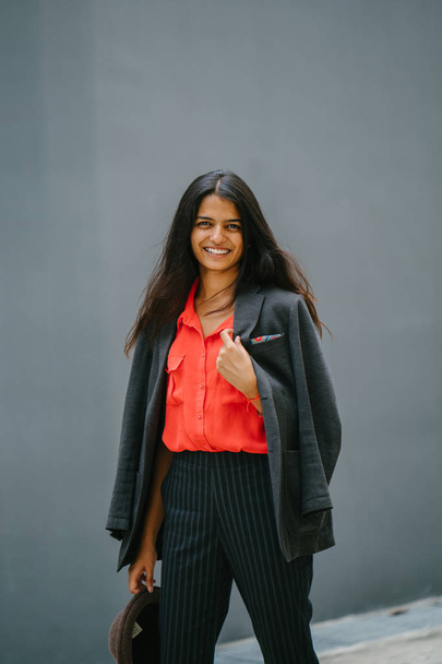 Portrait of a fashionable Indian Asian woman against a plain grey background. She is smiling and wearing a smart orange blouse and black pinstripe pants with a jacket draped over her shoulders. - Foto, Imagem