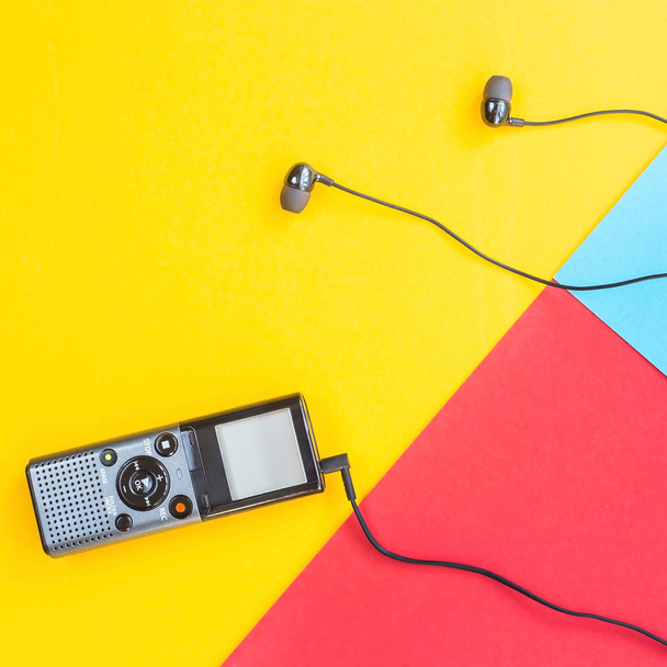 Black voice recorder with headphones on a combined yellow, blue and red background. Journalism concept. Equipment for work, interview and voice recording. Top view. Flat lay - 写真・画像