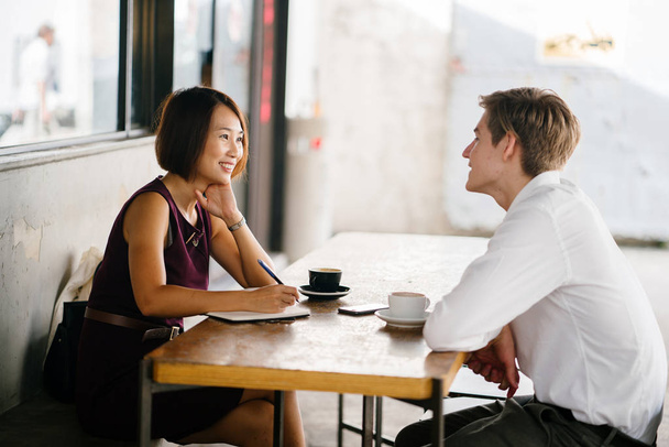 An Asian woman is interviewing a young Caucasian white man for an internship job in Asia. They are sitting at a desk in the shade outside in the day and talking over coffee. - Photo, Image