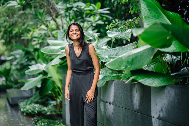 Candid portrait of a young Indian woman standing in a lush green garden. She's dressed in a professional grey smock jumpsuit and smiling. - Фото, изображение