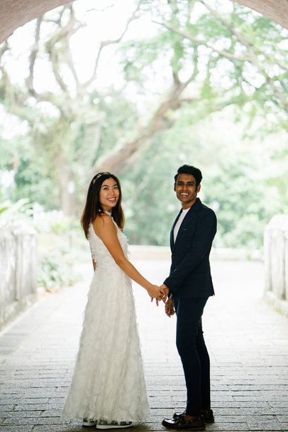  Indian groom holds the wife of his Chinese wife and stand side-by-side in a park. The interracial couple are taking wedding photographs under an arch in the day and are smiling. - Φωτογραφία, εικόνα