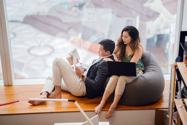 A Chinese Asian couple unwind over the weekend in their apartment. An attractive woman is working on her laptop on a beanbag and her boyfriend is reading a book and leaning against her. - Photo, image