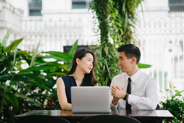 Portrait of two smiling business people (lawyers, consultants, etc) or couple smiling mid discussion over a laptop in an office with greenery in the background. They are focused on the laptop. - 写真・画像