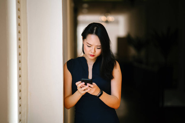 Retrato de una joven y atractiva mujer asiática china usando su teléfono inteligente cerca de una ventana en el día. Ella está usando un vestido azul oscuro profesional y sonriendo
. - Foto, Imagen