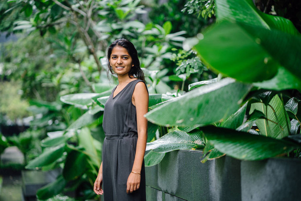Candid portrait of a young Indian woman standing in a lush green garden. She's dressed in a professional grey smock jumpsuit and smiling. - 写真・画像