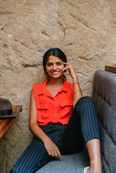 Portrait of a young and attractive Indian Asian woman smiling at the camera. She is seated and has a brown fedora cap . She is dressed in smart casual and is relaxed and cheerful. - Foto, Imagen