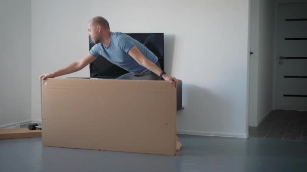 adult man is turning and lying on a floor big cardboard box in an room of apartment, cutting packing tape - Filmagem, Vídeo