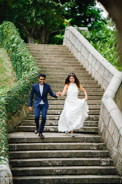 pair of interracial lovers run down stone steps in the park during the day. An Indian man is marrying a Chinese woman and they are running down the stairs in delight and joy. - Foto, Imagen