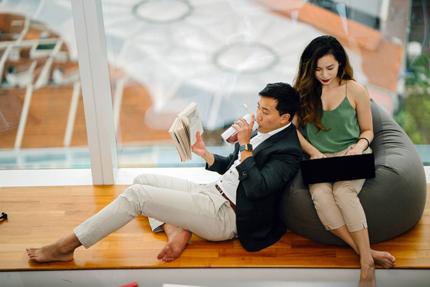 Китайская азиатская пара отдыхает на выходных в своей квартире. Привлекательная женщина работает над своим ноутбуком над бобовым мешком, а ее парень читает книгу и прислоняется к ней
. - Фото, изображение