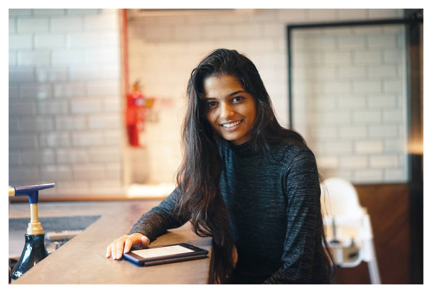 Портрет молодий привабливою Індійська жінка сидить за столом в кафе, випадково і природно посміхаючись від її планшетного ПК. - Фото, зображення