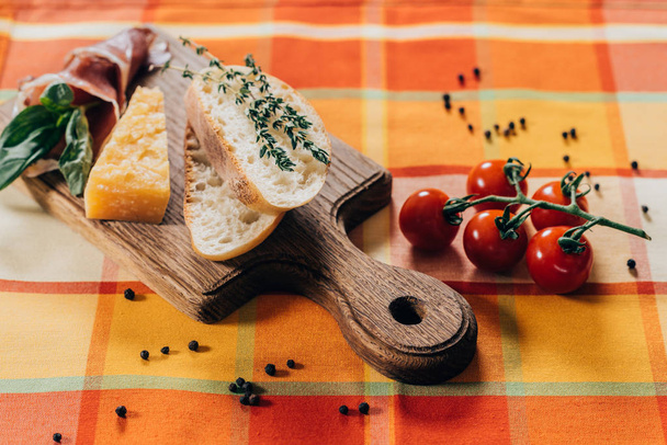 sliced baguette, meat and parmesan cheese on wooden cutting board and fresh cherry tomatoes on table  - Photo, image