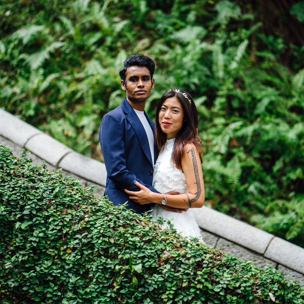 An attractive couple of different races stand together in park in day in Asia. They are taking wedding photographs. One is an Indian man, the other a Chinese woman. - Foto, afbeelding