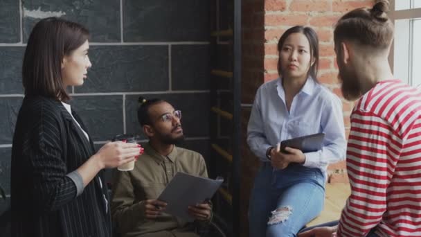 Group of multiracial colleagues disscussing startup project while standing near window in office with coffee and papers - Кадры, видео