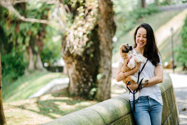 Portrait of a young, attractive and beautiful pan Asian woman with her pet shih tzuh dog in the day in a park with lots of greenery and plants in the background. - Photo, Image