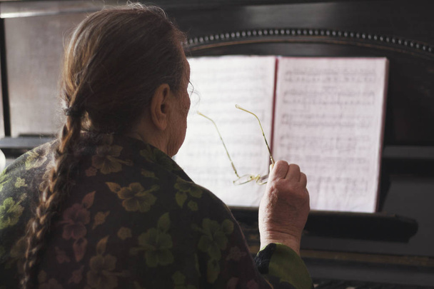 La anciana sentada frente a un piano y un cuaderno con gafas en las manos
 - Foto, Imagen