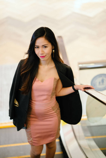 stylish and fashionable young Chinese Asian woman on an escalator. She is wearing a beautiful pink dress with a man's jacket draped over her shoulders and smiling. - Fotografie, Obrázek