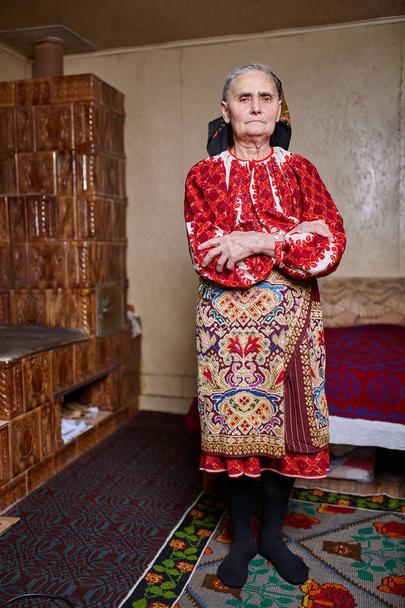 Old Romanian woman dressed in traditional costume standing at home - Foto, imagen