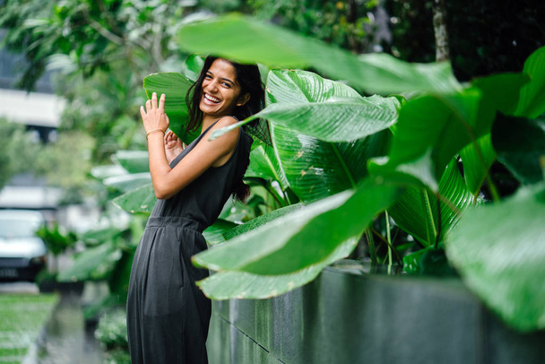 Candid portrait of a young Indian woman standing in a lush green garden. She's dressed in a professional grey smock jumpsuit and smiling. - Photo, image