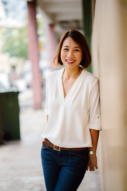A portrait of an attractive Chinese Asian woman leaning against a pastel yellow wall in Singapore, Asia. She is dressed in smart casual for the weekend and is smiling as she enjoys her day. - Фото, изображение