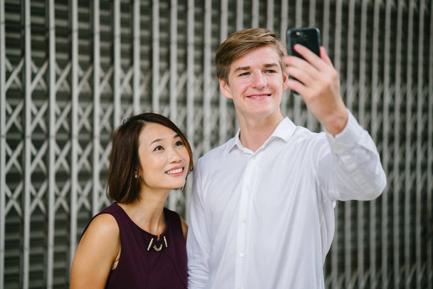gemengd-cultuur paar nemen een selfie in de dag tegen de metalen roosters van een gesloten Chinese winkel in Azië. Een jonge en aantrekkelijke witte man glimlacht naast een Chinese vrouw als ze hun foto nemen. - Foto, afbeelding