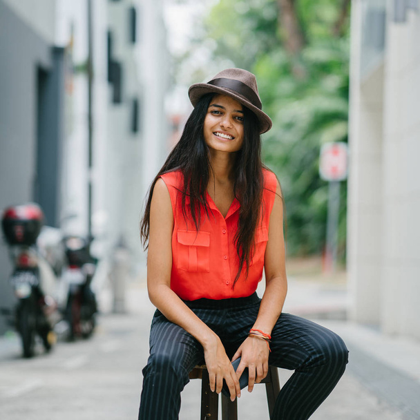 Portrait of an attractive, young and stylish Indian Asian woman sitting in a stool in an alley in Asia. She is smiling and dressed in smart casual and wearing a brown fedora hat. - Фото, изображение