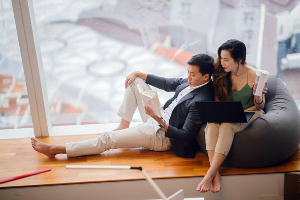 Китайская азиатская пара отдыхает на выходных в своей квартире. Привлекательная женщина работает над своим ноутбуком над бобовым мешком, а ее парень читает книгу и прислоняется к ней
. - Фото, изображение