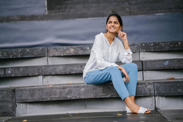 Candid portrait of a smiling attractive, young woman on grey steps . The Indian Asian lady is professional, relaxed and sitting in the bright sun. - Fotografie, Obrázek