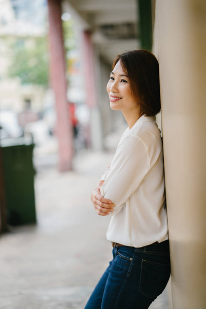 portrait of an attractive Chinese Asian woman leaning against a pastel yellow wall in Singapore, Asia. She is dressed in smart casual for the weekend and is smiling as she enjoys her day. - 写真・画像