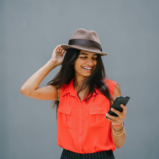 Portrait of a young and fashionable North Indian Desi Asian woman. She has a light complexion and is wearing an orange shirt, black pants and a fedora hat. She smiles as she uses her smartphone. - Foto, Bild