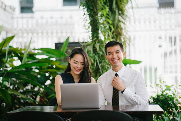 Portrait of two smiling business people (lawyers, consultants, etc) or couple smiling mid discussion over a laptop in an office with greenery in the background. They are focused on the laptop. - Foto, afbeelding