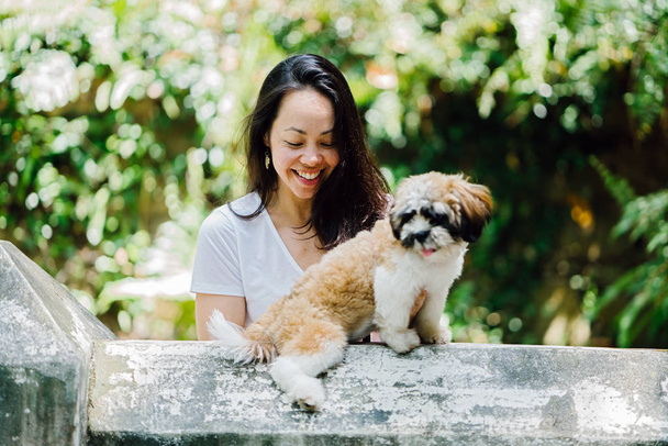 Portrait of a young, attractive and beautiful pan Asian woman with her pet shih tzuh dog in the day in a park with lots of greenery and plants in the background. - Foto, imagen
