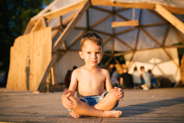 Theme is yoga and children. Caucasian Boy child sitting barefoot cross-legged in lotus position on wooden floor the background of gathering place of people. In summer sunset. Dressed in denim shorts - Photo, Image