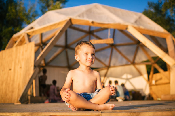 Le thème est yoga et enfants. Enfant garçon caucasien assis pieds nus jambes croisées en position lotus sur le sol en bois le fond de lieu de rassemblement des gens. Au coucher du soleil d'été. Vêtu de shorts en denim
 - Photo, image