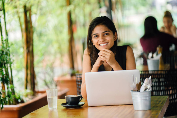 Candid portrait of an attractive and young Indian Asian professional woman work on her laptop in a trendy cafe or coworking space with teal wooden furniture. She is smiling and looking away. - Photo, image