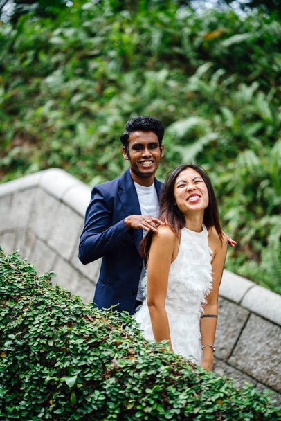 An attractive couple of different races stand together in park in day in Asia. They are taking wedding photographs. One is an Indian man, the other a Chinese woman. - Photo, image