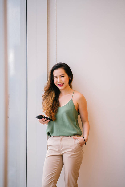 Portrait of an attractive, young Singaporean woman in her comfortable loft apartment. She is standing next to a window and smiling contentedly. - Foto, Bild