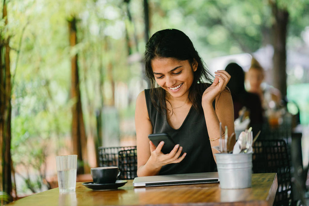 Openhartig portret van een aantrekkelijke, jonge Indiase vrouw zitten in een cafe of coworking space met smartphone. op een zonnige dag. Er zijn weelderige, groene planten op de achtergrond. - Foto, afbeelding