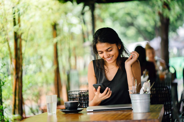Candid portrait of an attractive, young Indian woman sitting in a cafe or coworking space with smartphone. on a sunny day. There are lush, green plants in the background. - Foto, imagen
