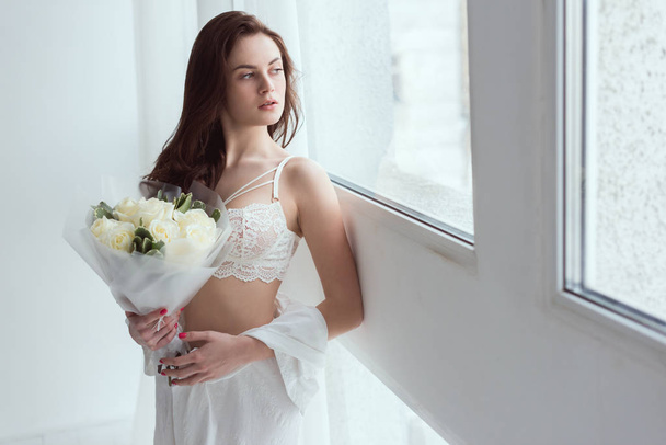 young pensive woman in underwear with bouquet of roses looking out window - Foto, immagini