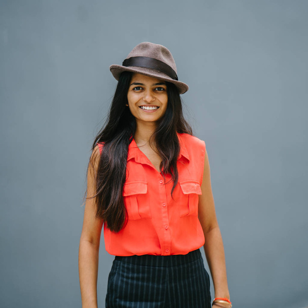 Portrait of a young, attractive Indian Asian fashion student. She is smiling and standing in the sun against a plain grey blue background. - Foto, Imagem