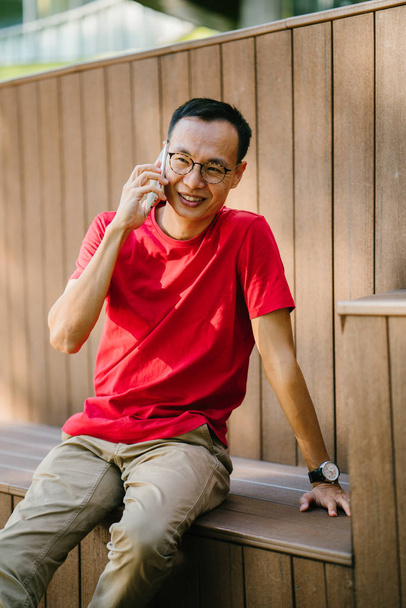 middle-aged Asian man talking on his phone in a park. He is sitting on a wooden structure and smiles as he types on his phone at sunset. He is dressed comfortably in a tee shirt and loose pants. - Fotoğraf, Görsel