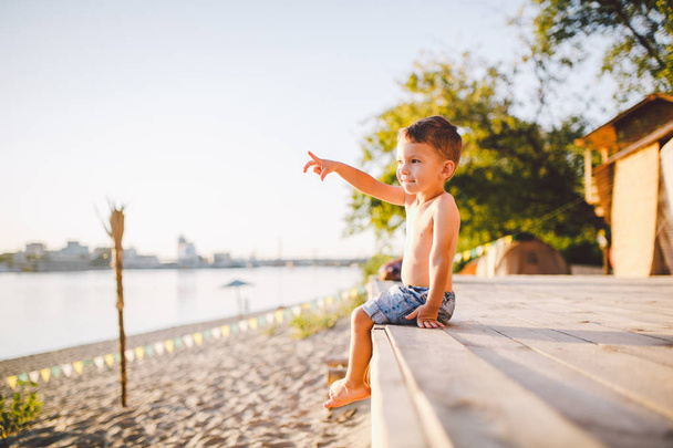 Thema ist ein Kinder- und Sommerurlaub am Strand. Ein kleiner kaukasischer Junge sitzt seitlich auf einem Holzsteg und zeigt seine Hand auf einen Sandstrand und einen Teich, einen Fluss. mit nackten Beinen in blauen Jeanshosen - Foto, Bild