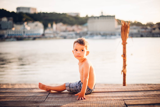 Темою є дитина і літній пляжний відпочинок. Маленький хлопчик кавказьких вбік сидить на дерев'яний причал і дивиться на камеру на піщаний пляж та ставок, річка. З голими ногами у синій джинсового шорти - Фото, зображення