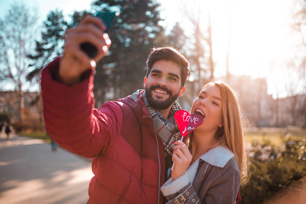 Güzel genç çift birlikte parkta öpüşme ve kalp şeklinde bir lolipop tutan, tadını seviyorum. Çift alarak selfie. - Fotoğraf, Görsel