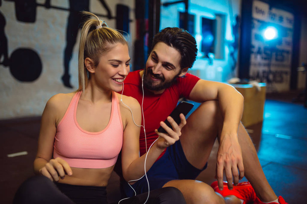 Young sporty couple using mobile phone in gym. Friends listening to the music in gym. - Photo, Image