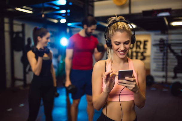 Portrait of young sportswoman with smartphone listening to music in gym. - Photo, Image
