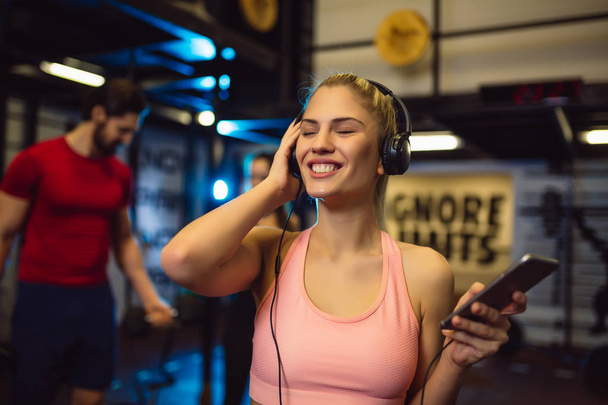 Portrait of young sportswoman with smartphone listening to music in gym. - 写真・画像